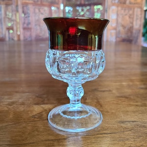 May include: A clear glass goblet with a ruby red rim and a decorative, patterned bowl. The goblet has a delicate stem and a circular base. The rim has a gold-colored band. The glass is likely vintage or antique.