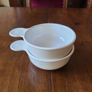 May include: Two off-white soup bowls with handles are stacked on a wooden table. The bowls are round with a slightly flared rim and a single handle. The background includes a wooden table and a dark wooden cabinet.