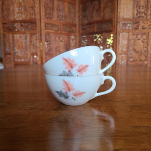 May include: Two white teacups stacked on a wooden table. Each cup features a delicate design of pink and gray leaves. The cups have a classic shape with a curved handle. The background includes a wooden folding screen.