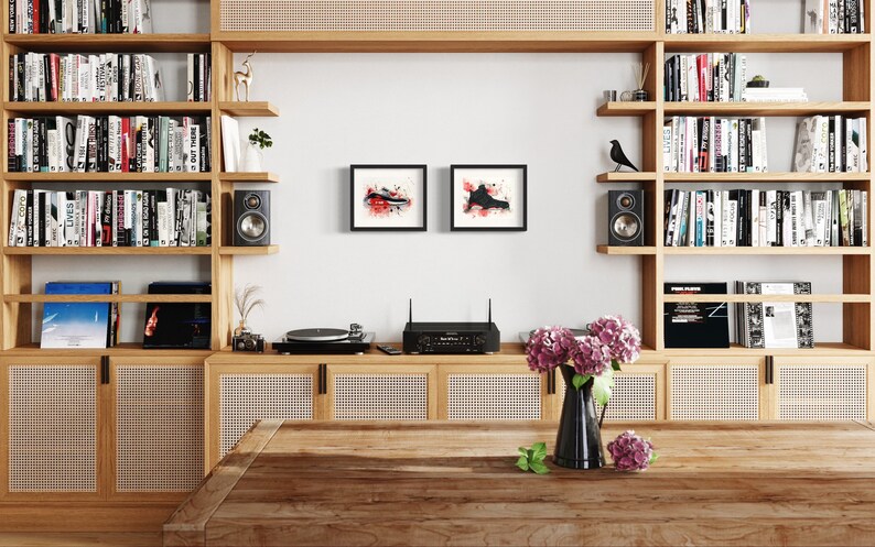 May include: A wooden bookshelf with a record player, amplifier, and speakers. The bookshelf is filled with books and has a woven cane panel on the bottom. There are two black framed abstract paintings on the wall above the record player.