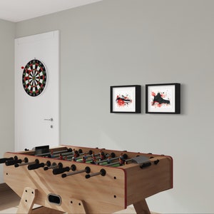 May include: A wooden foosball table with black handles and a red trim. There is a dartboard on the wall behind the table, and two framed black and white prints with red accents on the wall.