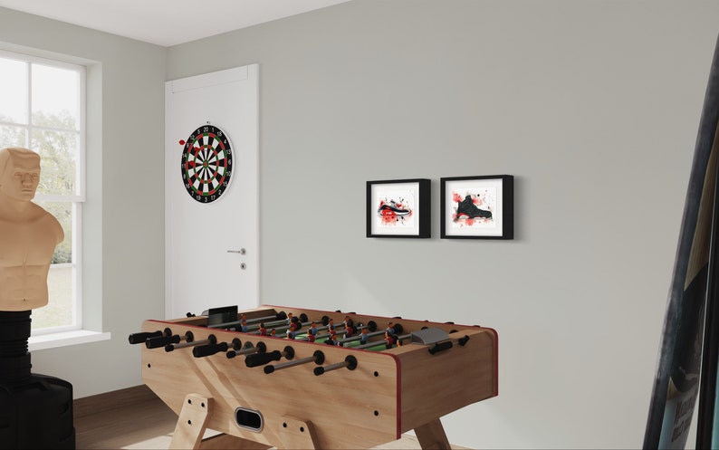May include: A wooden foosball table with black handles and a red trim. There is a dartboard on the wall behind the table, and two framed black and white prints with red accents on the wall.