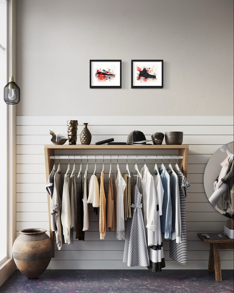 May include: A white wall with a wooden shelf holding a clothing rack with various clothing items hanging on it. There are two framed black and white watercolor paintings of sneakers on the wall above the shelf.