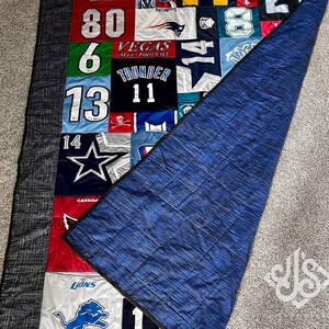 K&ouml;nnte beinhalten: Eine blaue und graue Steppdecke mit Patchwork-Muster, das verschiedene Sportteam-Logos und Nummern zeigt. Die Steppdecke ist in der H&auml;lfte gefaltet und zeigt eine blaue R&uuml;ckseite.