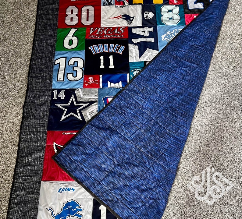 K&ouml;nnte beinhalten: Eine blaue und graue Steppdecke mit Patchwork-Muster, das verschiedene Sportteam-Logos und Nummern zeigt. Die Steppdecke ist in der H&auml;lfte gefaltet und zeigt eine blaue R&uuml;ckseite.