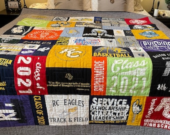 May include: A handmade quilt made from repurposed t-shirts. The quilt features a patchwork design with various school-related graphics, including the text "Class of 2021", "RC Eagles", and "Track & Field". The quilt is displayed on a bed.