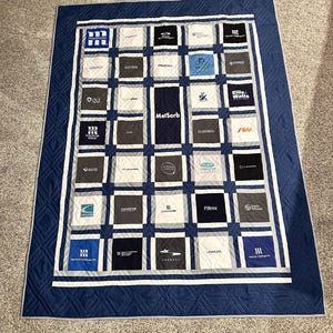 May include: A handmade quilt with a grid pattern, featuring squares in white, gray, and navy blue. Each square displays a company logo. The quilt has a navy blue border with white accents.