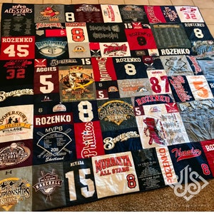 K&ouml;nnte beinhalten: Eine Patchwork-Decke aus verschiedenen T-Shirts mit Baseball- und Sportmotiven. Die Decke besteht aus Quadraten in verschiedenen Farben und Mustern, darunter Rot, Wei&szlig;, Blau und Schwarz. Die Decke zeigt verschiedene Logos und Texte, darunter "World Series", "Superbowl Saturday", "Brewers", "Phillies", "Murrieta All Stars", "Aggies", "Thunder", "Rozenko", "MVPB" und "Cooperstown All Star Village".