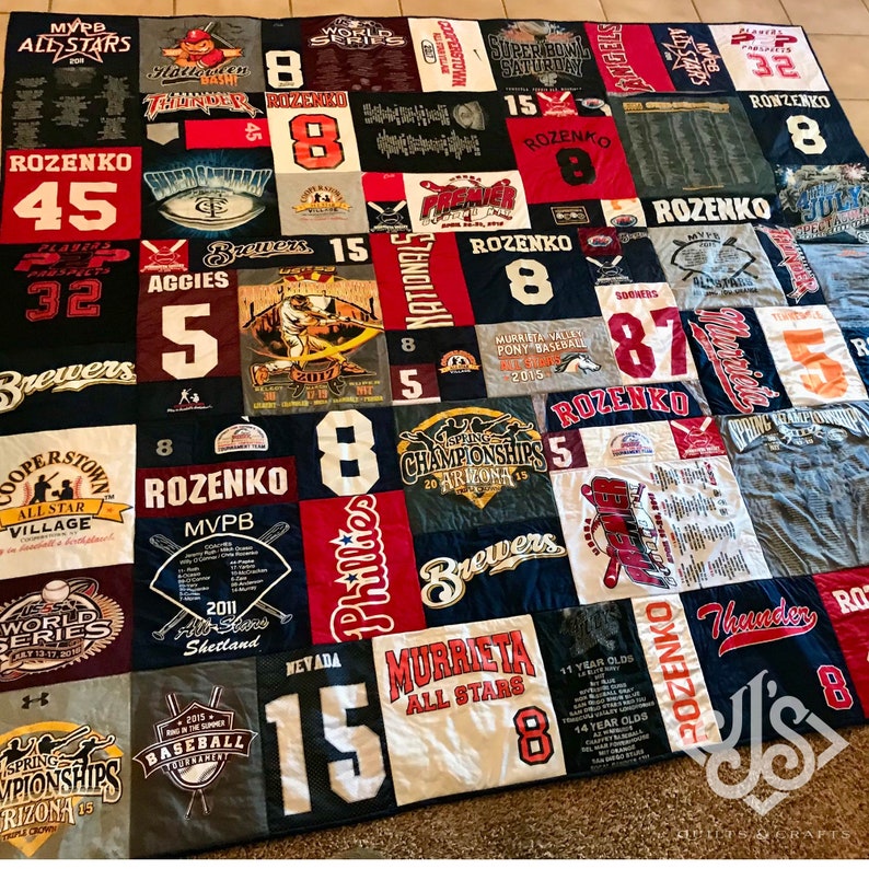 K&ouml;nnte beinhalten: Eine Patchwork-Decke aus verschiedenen T-Shirts mit Baseball- und Sportmotiven. Die Decke besteht aus Quadraten in verschiedenen Farben und Mustern, darunter Rot, Wei&szlig;, Blau und Schwarz. Die Decke zeigt verschiedene Logos und Texte, darunter "World Series", "Superbowl Saturday", "Brewers", "Phillies", "Murrieta All Stars", "Aggies", "Thunder", "Rozenko", "MVPB" und "Cooperstown All Star Village".