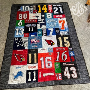 K&ouml;nnte beinhalten: Eine Patchwork-Decke aus verschiedenen Sporttrikots. Die Decke zeigt eine Vielzahl von Teamlogos und Nummern, darunter die Cardinals, Panthers, Saints, Gators und viele mehr. Die Decke ist mit einem schwarzen und grauen Stoffrand gefertigt.