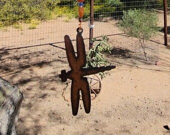 May include: A rusty metal dragonfly wind chime with colorful glass beads hanging from a chain. The wind chime is hanging from a tree branch.