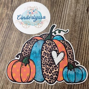 May include: A sticker featuring a large pumpkin design with a leopard print pattern, orange polka dots, and a blue section. A white heart is cut out of the pumpkin. A smaller sticker with the text "Cinderlyssa" and a floral carriage design is also present.