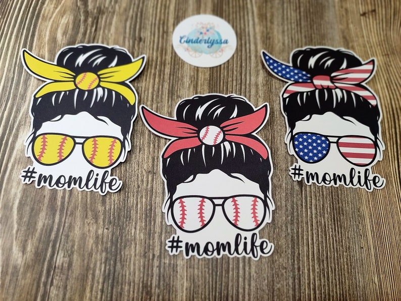 Peut inclure: Trois autocollants en vinyle repr&eacute;sentant une femme de dessin anim&eacute; avec un chignon d&eacute;sordonn&eacute;, des lunettes de soleil et un bandeau. Les bandeaux sont jaunes, rouges et rouge, blanc et bleu. Les lunettes de soleil sont d&eacute;cor&eacute;es d'un motif de baseball. Chaque autocollant porte le texte "#momlife" sous le dessin anim&eacute;.