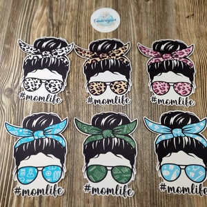 Peut inclure: Six autocollants en vinyle diff&eacute;rents repr&eacute;sentant une femme de dessin anim&eacute; avec un chignon d&eacute;sordonn&eacute;, des lunettes de soleil et un bandana. Chaque autocollant porte le texte "#momlife" sous l'image.