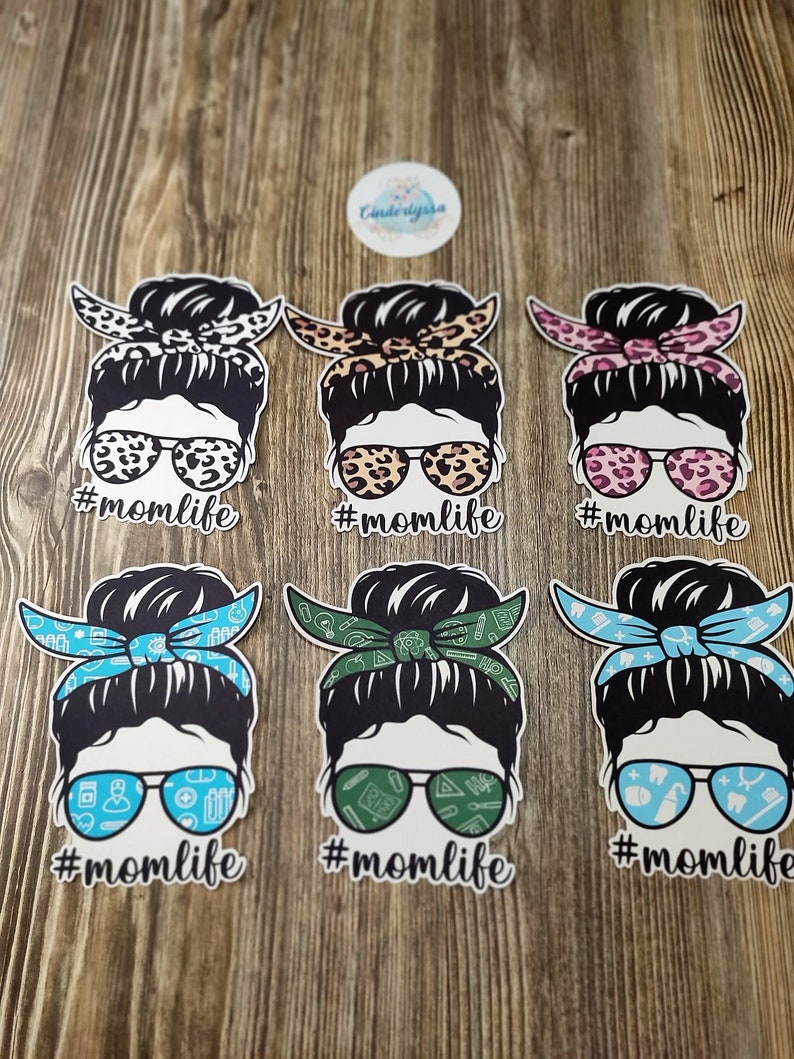 Peut inclure: Six autocollants en vinyle diff&eacute;rents repr&eacute;sentant une femme de dessin anim&eacute; avec un chignon d&eacute;sordonn&eacute;, des lunettes de soleil et un bandana. Chaque autocollant porte le texte "#momlife" sous l'image.