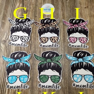 Peut inclure: Six designs diff&eacute;rents d'une femme avec un chignon d&eacute;sordonn&eacute; portant des lunettes de soleil et un bandana. Les bandanas sont de motifs diff&eacute;rents, notamment un imprim&eacute; l&eacute;opard, des fleurs roses, des symboles m&eacute;dicaux verts et bleus et des symboles dentaires bleus et blancs. Chaque design comporte le texte "#momlife" en dessous.