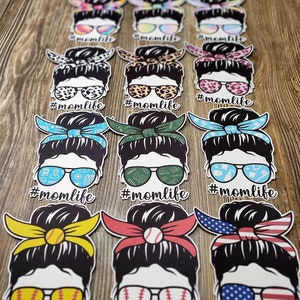 Peut inclure: Un ensemble de 12 autocollants en vinyle color&eacute;s repr&eacute;sentant une femme de dessin anim&eacute; avec un chignon d&eacute;sordonn&eacute;, des lunettes de soleil et un bandana. Chaque autocollant porte le texte "#momlife" sous l'image. Les bandanas sont de motifs diff&eacute;rents, notamment des imprim&eacute;s l&eacute;opard, floraux et &agrave; carreaux.