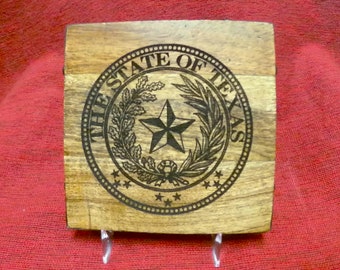 May include: A wooden coaster with the State of Texas seal engraved on it. The seal features a five-pointed star, a wreath of olive branches, and the words "The State of Texas".