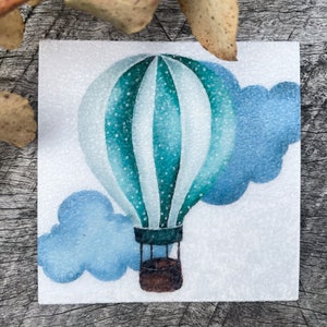 Puede incluir: Imagen cuadrada, estilo acuarela, que presenta un globo aerostático en tonos turquesa y blanco, con una cesta marrón. Una nube azul está pintada debajo del globo. La imagen está sobre un fondo blanco, con una superficie de madera en el fondo.