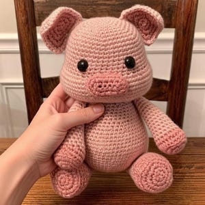 May include: A handmade, light pink crochet pig stuffed animal with black safety eyes and a detailed snout. The pig has floppy ears and is held in a person's hand. The toy is made of soft yarn and is sitting on a wooden surface.
