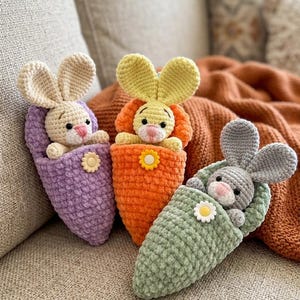 May include: Three crocheted bunny dolls in purple, orange, and green cocoons. Each bunny has large ears and a flower accent. The dolls are resting on a neutral-toned couch.