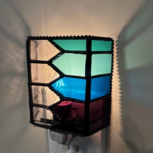 May include: A stained glass night light with a black metal frame. The glass panels are teal, blue, and purple. The night light is plugged into a white electrical outlet on a light-colored wall.