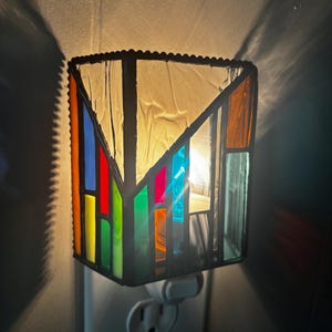 May include: A rectangular stained glass night light plugged into a wall outlet. The lamp features a geometric pattern with panels of clear, orange, blue, red, and green glass held together by dark metal solder lines, emitting a warm internal glow.