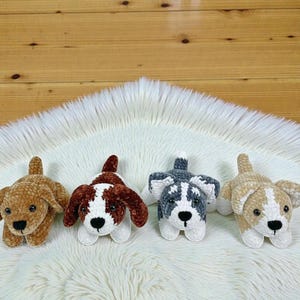 May include: Four handmade crochet dog toys in various colors: tan, brown and white, gray and white, and tan and white. The toys are lying on a white, fluffy surface. Each dog has a distinct color pattern and facial features.