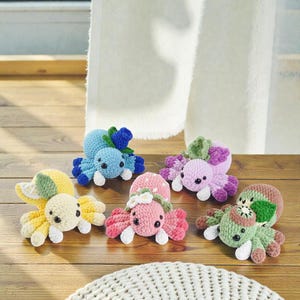 Puede incluir: Seis juguetes de pulpo de ganchillo hechos a mano en tonos amarillo, azul, rosa y verde, con detalles blancos. Cada pulpo tiene un diseño único, incluyendo detalles de flores y hojas. Están dispuestos sobre una superficie de madera.