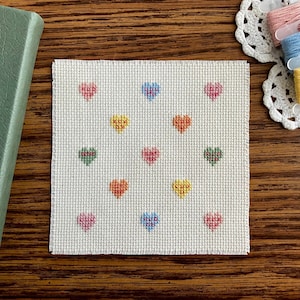 Puede incluir: Un cuadrado de tela de punto de cruz blanquecina con una cuadrícula de corazones coloridos en rosa, amarillo, verde y azul. Un libro verde y hilo de bordar rosa, azul y amarillo también son visibles.