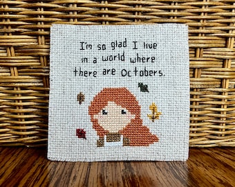 May include: Cross-stitch embroidery with a quote: "I'm so glad I live in a world where there are Octobers."  A cartoon girl with red hair is in the center of the design, surrounded by autumn leaves.