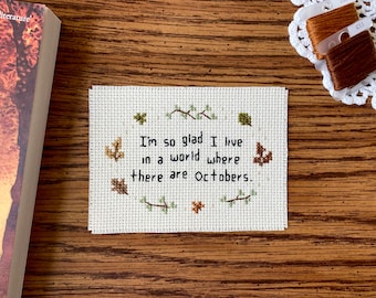 May include: A white cross-stitch embroidery with the quote "I'm so glad I live in a world where there are Octobers." surrounded by brown and green embroidered leaves.