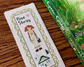 May include: A white cross-stitch bookmark with a green border and a floral design. The bookmark features a cartoon image of Anne Shirley from the book "Anne of Green Gables" with the text "Anne Shirley" above her.