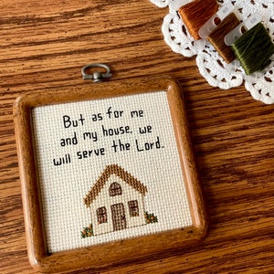 May include: A framed cross-stitch with a white background and brown wooden frame. The cross-stitch depicts a small house with a brown roof and a white door. The text "But as for me and my house, we will serve the Lord." is stitched in black thread.