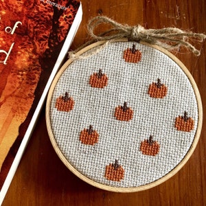 May include: A round, wooden-framed embroidery hoop with a white fabric background and a repeating pattern of orange pumpkins.