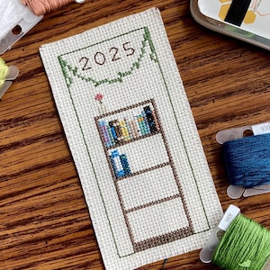 Pode incluir: Um marcador de página branco de ponto cruz com um design de estante de livros e o ano 2025 bordado em fio marrom. O marcador de página é cercado por fio verde e tem uma pequena flor vermelha bordada na parte superior.