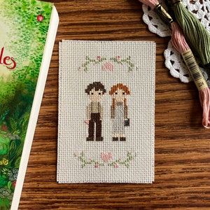 May include: Cross-stitch embroidery depicting two figures, a boy and a girl, standing together. They are surrounded by a wreath of green leaves and pink flowers. The background is white.