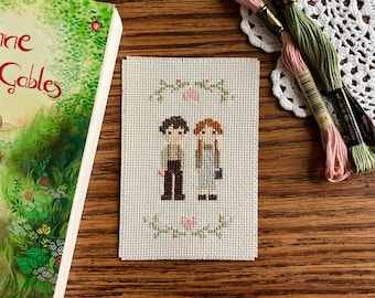 May include: Cross-stitch embroidery of two figures, a boy and a girl, standing side-by-side. They are surrounded by a wreath of green leaves and pink flowers. The background is white.