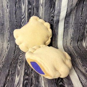May include: Two plush pillows shaped like hand pies. The pillows are beige with a brown crust and a purple filling.
