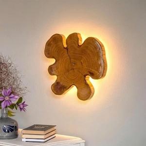 May include: A wall-mounted light fixture crafted from a natural wood slice with a visible grain pattern. The irregular, organic shape of the wood is illuminated from behind, casting a warm, soft glow against the wall surface behind it.