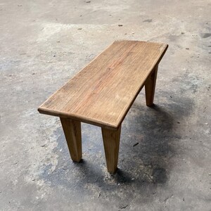 May include: A small, rectangular wooden bench with a natural finish. The bench has a simple design with four sturdy legs and a flat seat. The wood grain is visible, adding to its rustic charm. The bench is suitable for indoor or outdoor use.