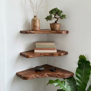 May include: Three triangular corner shelves made of dark brown wood with natural live edges. The top shelf holds a tan ceramic vase with dried grass and a small bonsai tree. The middle shelf holds a stack of three books. The bottom shelf holds a dark stone.