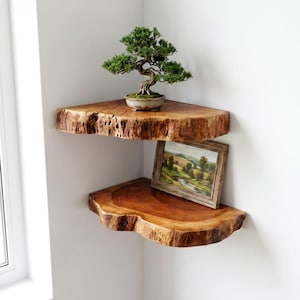 Może przedstawiać: Dwie półki narożne wykonane z plastrów naturalnego drewna z surowymi krawędziami. Górna półka mieści małe zielone drzewko bonsai w ceramicznej doniczce. Dolna półka prezentuje oprawiony obraz przedstawiający krajobraz z zielonymi wzgórzami.