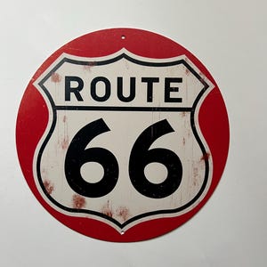 以下が含まれることがあります： 赤と白の丸いルート66の標識。黒い文字で「ROUTE 66」と数字の「66」が描かれています。標識は、使い古されたようなヴィンテージ感があります。