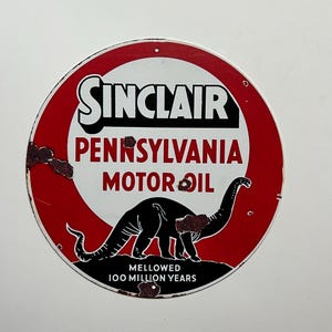 Puede incluir: Una señal de metal circular roja y blanca con el logotipo de Sinclair y el texto "Pennsylvania Motor Oil". Un dinosaurio negro aparece en la señal con el texto "Mellowed 100 Million Years".