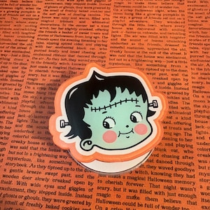 May include: A circular phone grip featuring a cartoon illustration of a green-skinned character with black hair, a stitched forehead, and silver bolts on the sides of the head. The character has large eyes, pink cheeks, and a small smile.