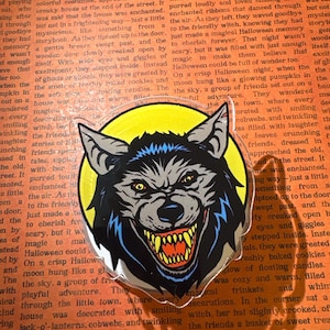 May include: A clear acrylic charm featuring a graphic illustration of a snarling wolf head with grey fur, pointed ears, and yellow eyes. The wolf has an open mouth revealing sharp yellow teeth and a red tongue, set against a bright yellow circular background.