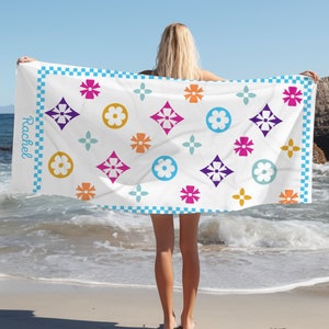 May include: A white beach towel with a colorful floral and geometric pattern. The towel features a blue checkered border and the name "Rachel" printed in script. The design includes flowers and diamond shapes in shades of pink, orange, purple, and blue.