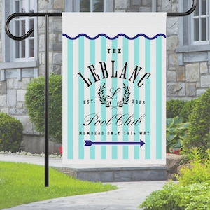 May include: A personalized garden flag with a teal and white striped background. The flag features the text "THE LEBLANC POOL CLUB" with a laurel wreath and an arrow pointing right. The flag is displayed on a black metal stand.