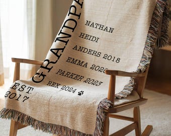 May include: A beige woven throw blanket draped over a wooden rocking chair. The blanket features the word "GRANDPA" vertically, along with names and dates in black text. The blanket has a fringed edge and the text "EST 2017".